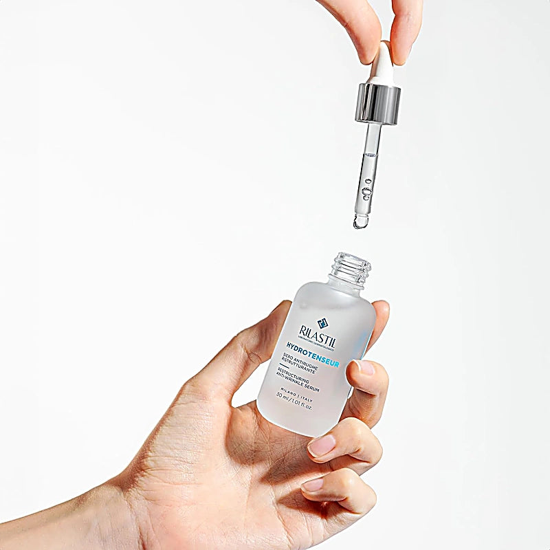 Hand holding a bottle of Rilastil Hydro Intense serum with a dropper on a white background